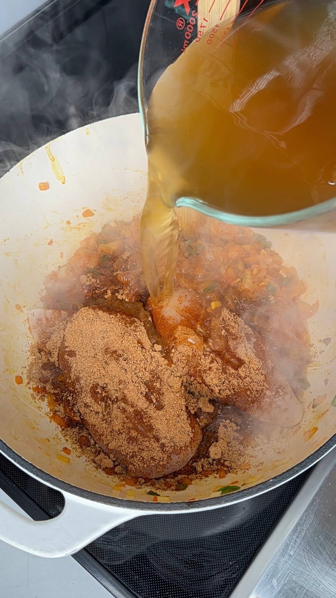 Liquid being poured from a glass measuring cup into a white pot containing raw chicken breasts covered in brown seasoning and sautéed vegetables.