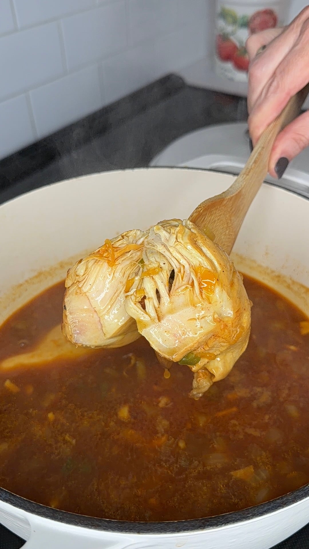 A wooden spoon lifts shredded, cooked chicken out of a pot of steaming, reddish-brown broth.