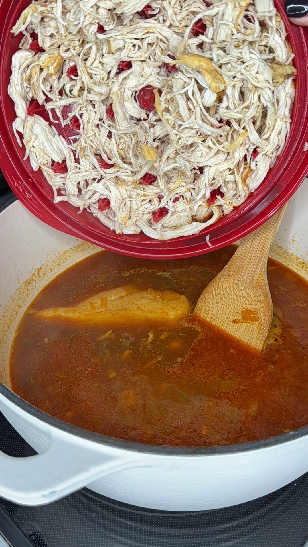 Shredded chicken in a red lid being poured into a white pot of simmering soup with a wooden spoon.