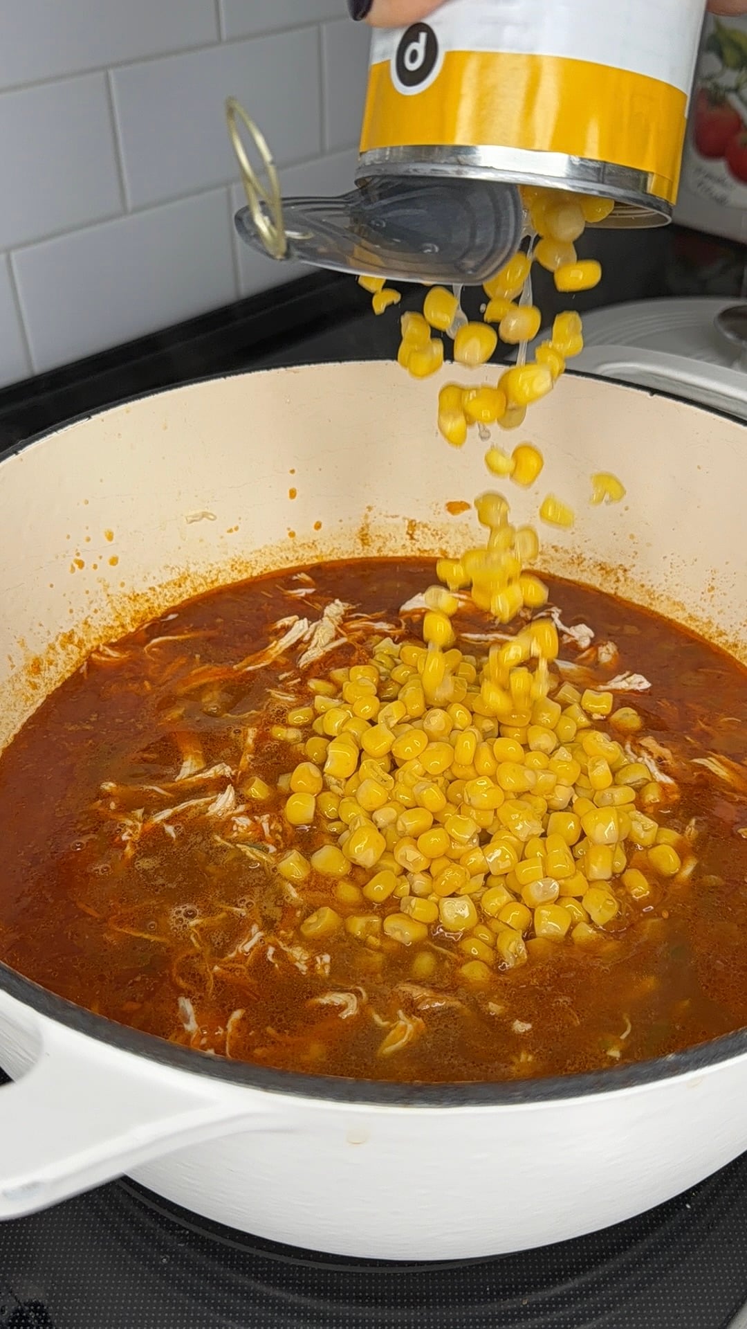 Canned corn being poured from a yellow and white can into a pot of red soup with shredded chicken.