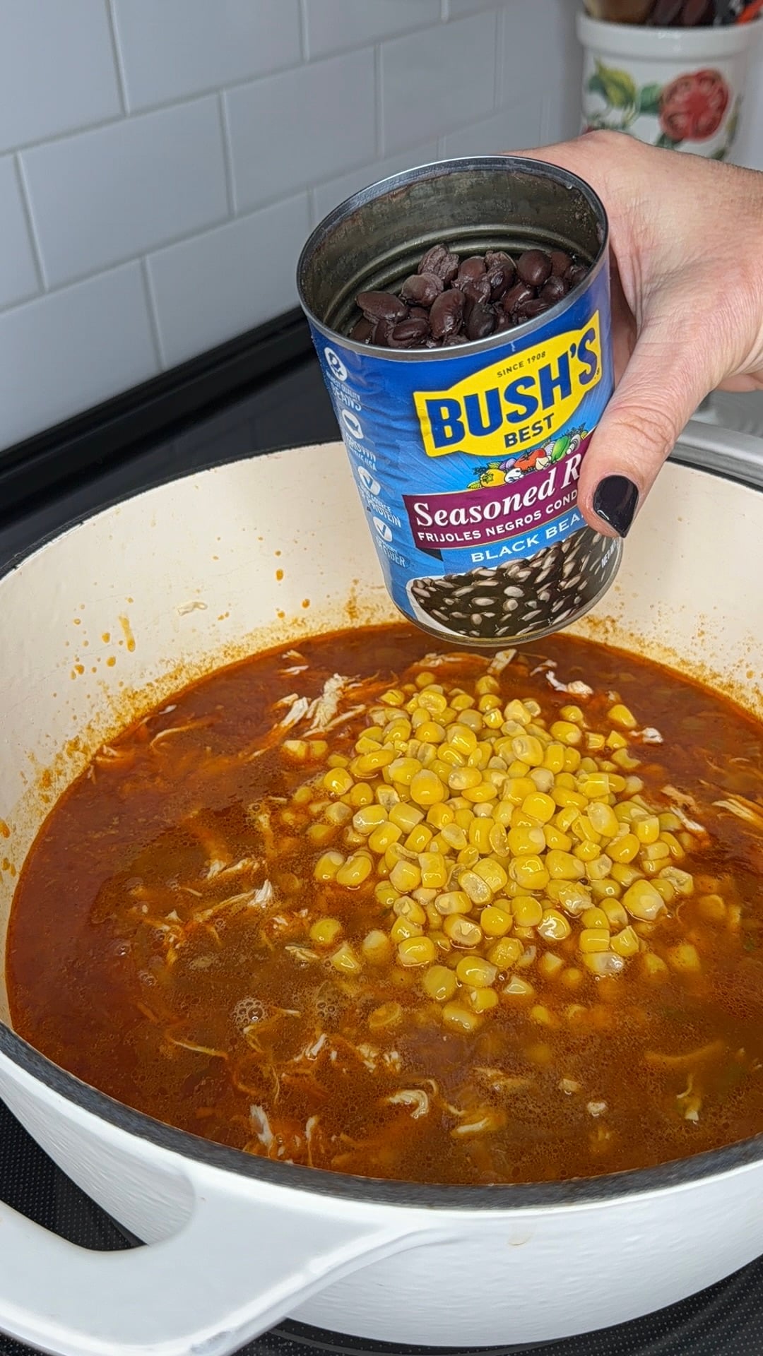 A hand pours a can of Bushs Best black beans into a pot of soup containing corn and shredded chicken.