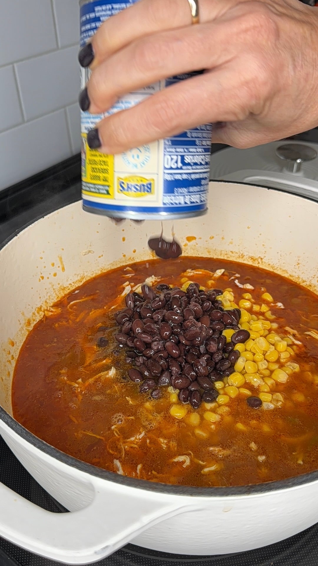 A hand pours a can of Bushs black beans into a pot of simmering soup containing corn and shredded chicken.