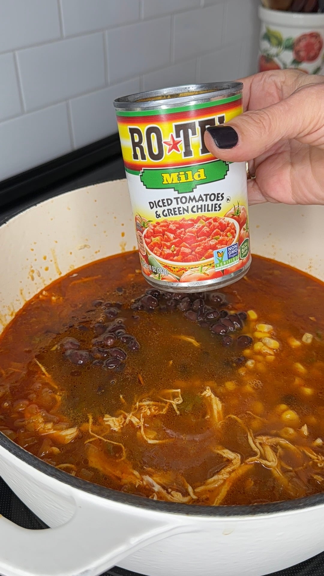 A hand pours a can of Ro-Tel Mild diced tomatoes and green chilies into a pot of soup containing shredded chicken, black beans, and corn.