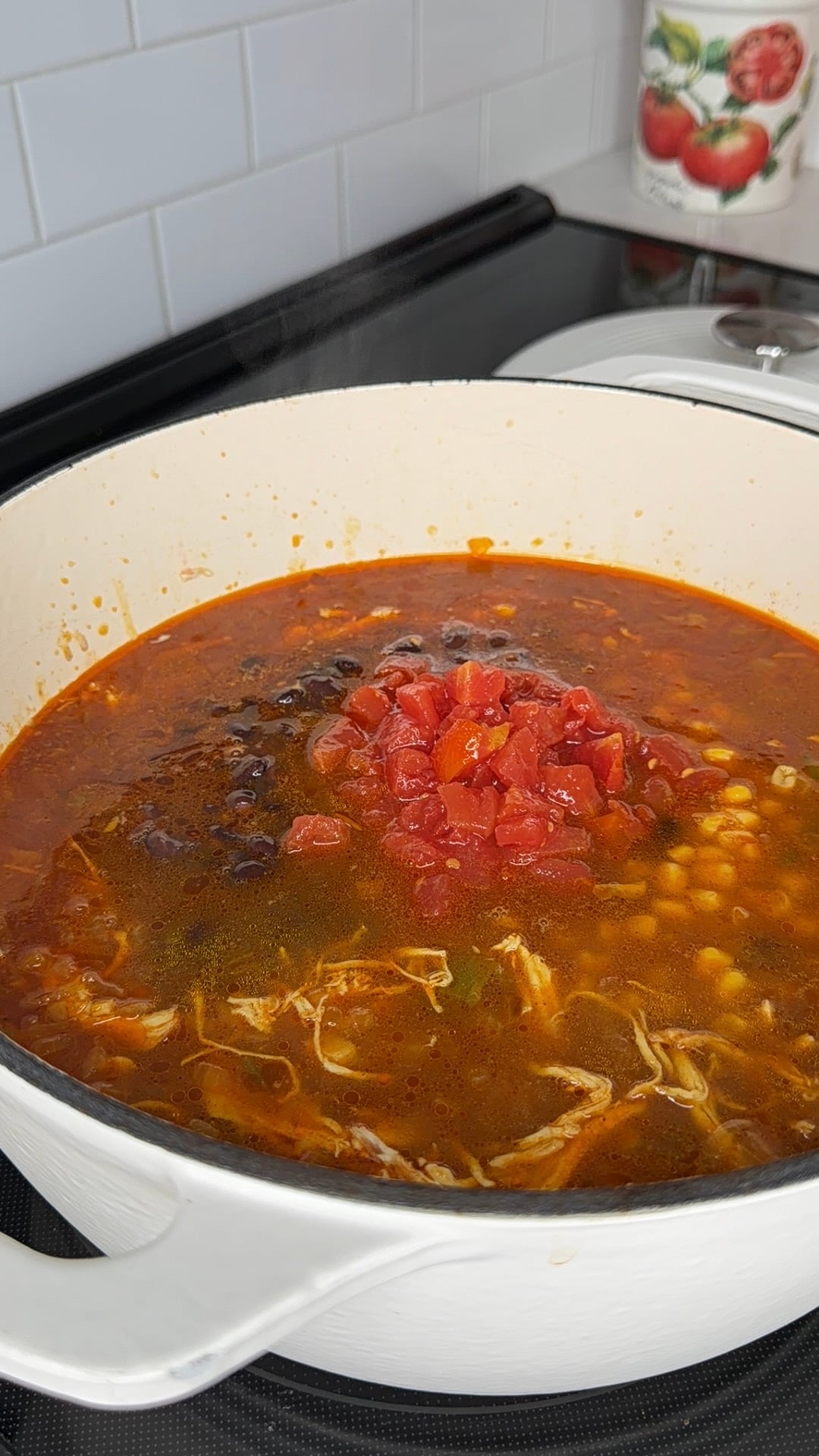 A large pot of soup on a stovetop, containing shredded chicken, black beans, corn, and a pile of diced tomatoes.