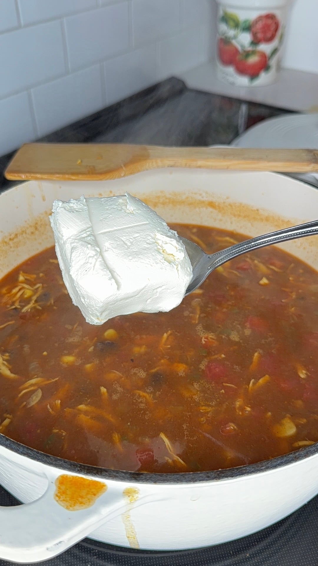 A spoonful of cream cheese held over a pot of tomato-based soup with shredded chicken.