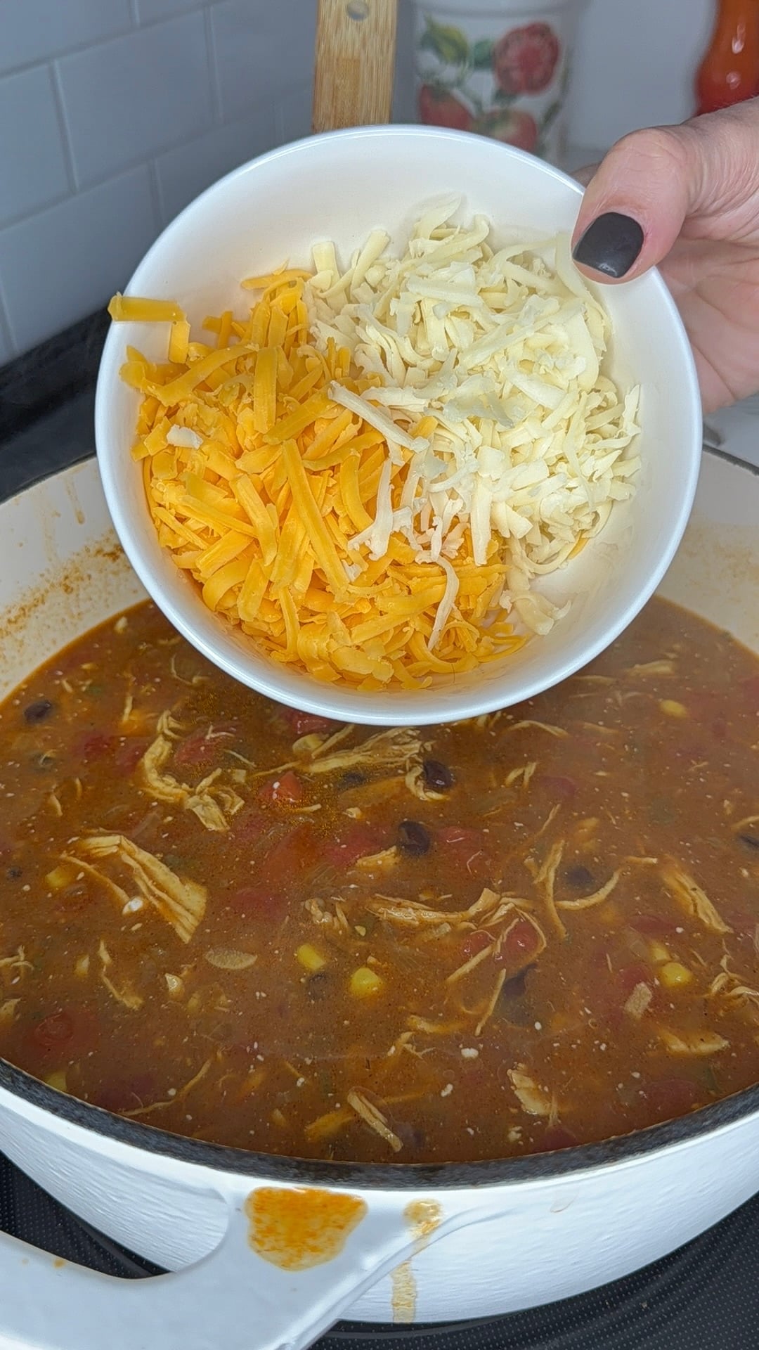 A hand holds a white bowl of shredded cheddar and white cheese over a pot of chicken and bean soup.