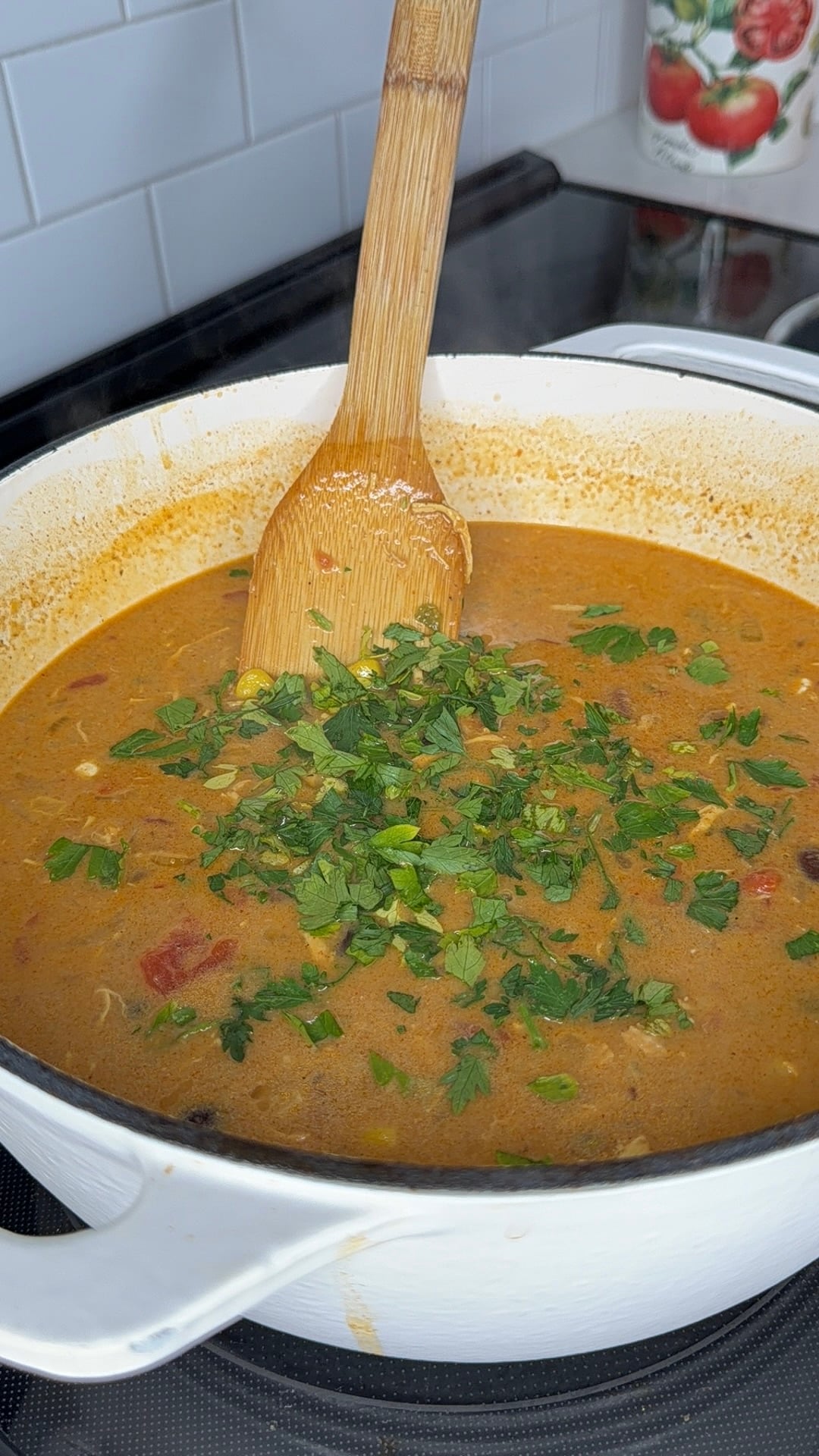 A bamboo spoon stirs a creamy, orange-colored soup topped with fresh chopped parsley in a white pot.