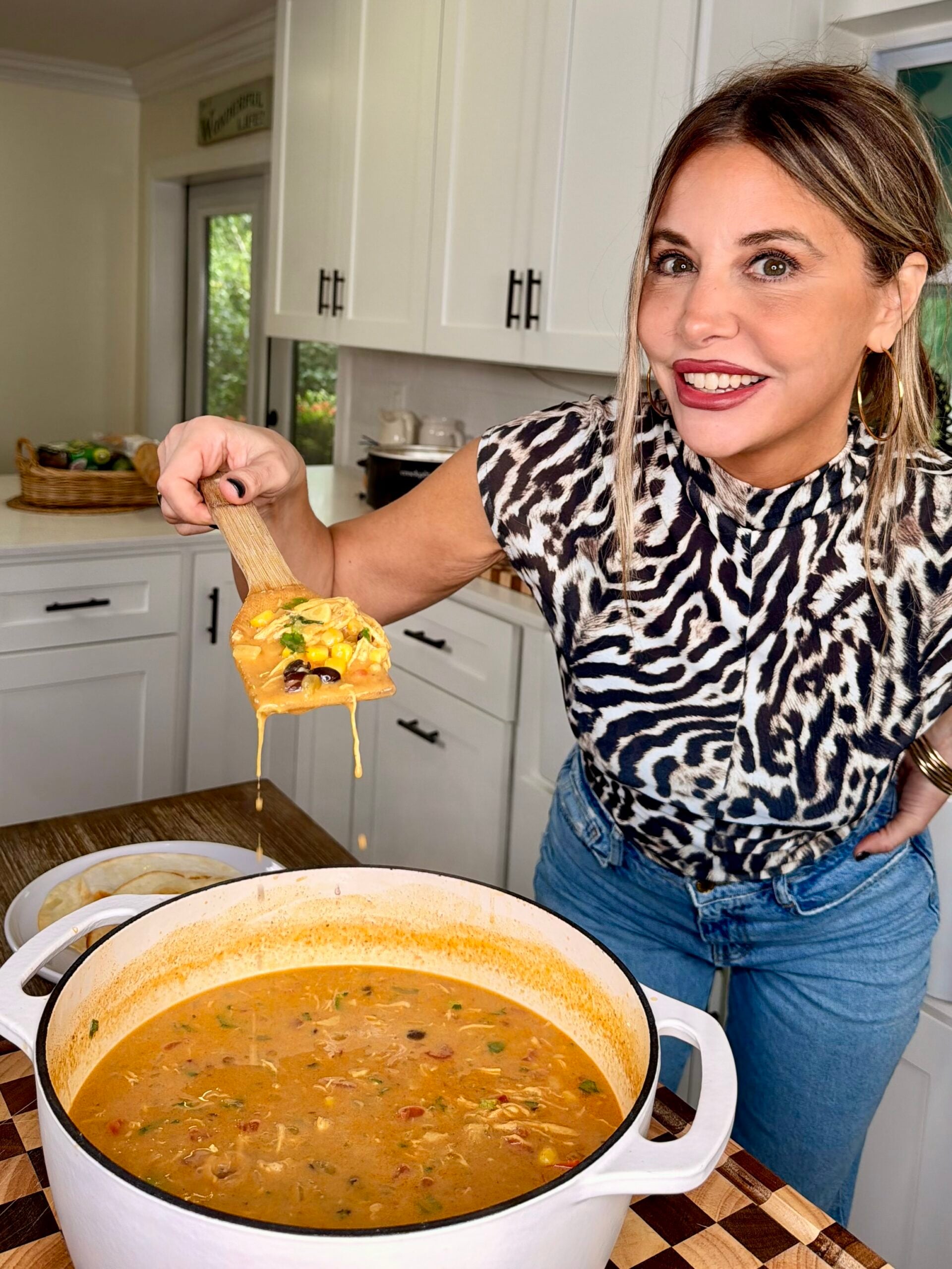 A woman holding a wooden spoon with chicken tortilla soup over a large white pot.
