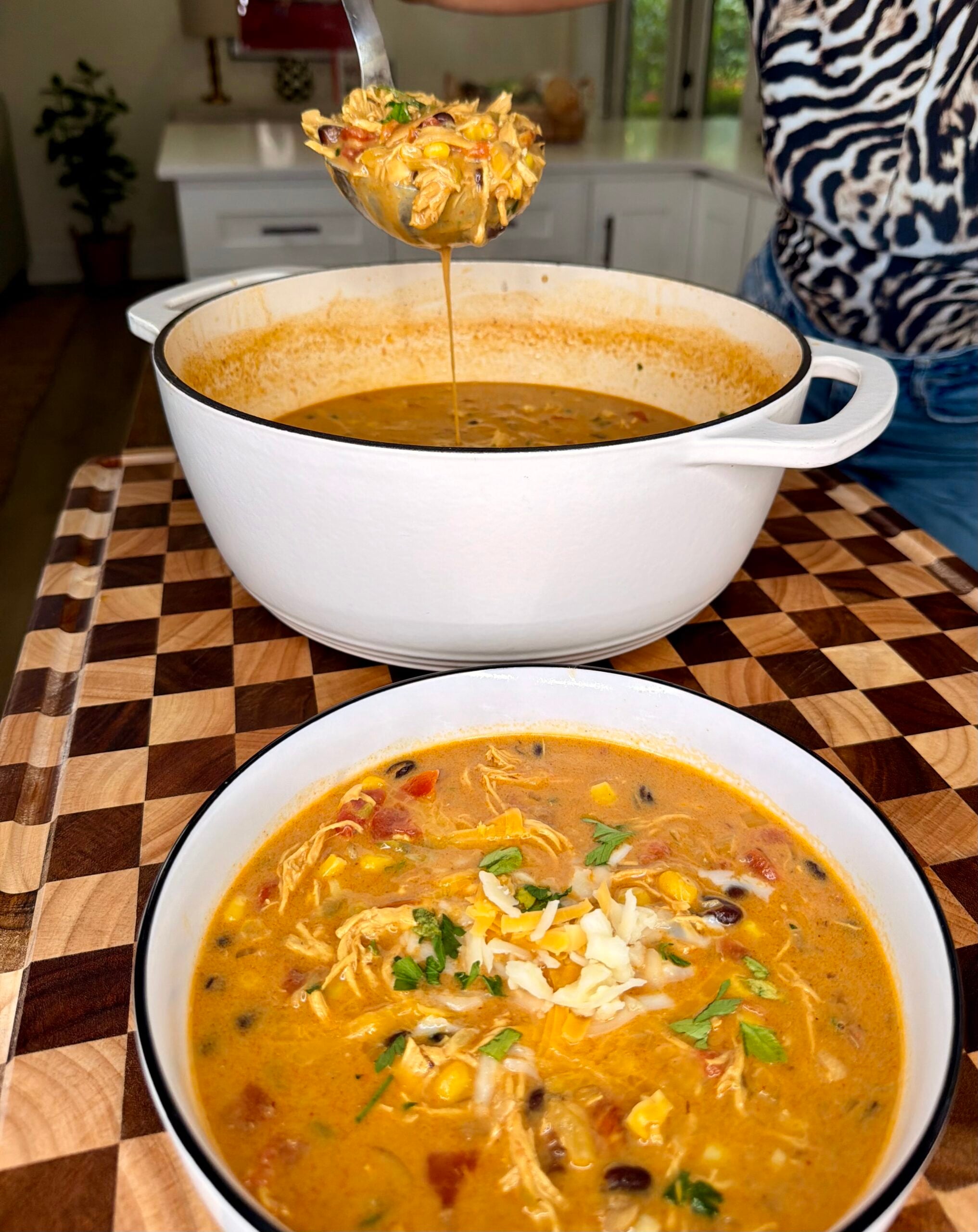 A ladle lifts chicken tortilla soup from a large white pot into a bowl topped with shredded cheese and cilantro.