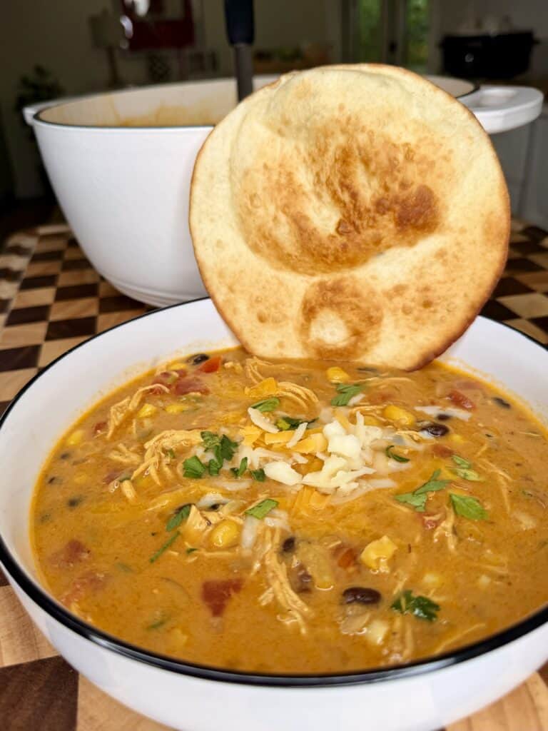 A bowl of chicken tortilla soup topped with shredded cheese and cilantro, served with a round crispy flatbread.