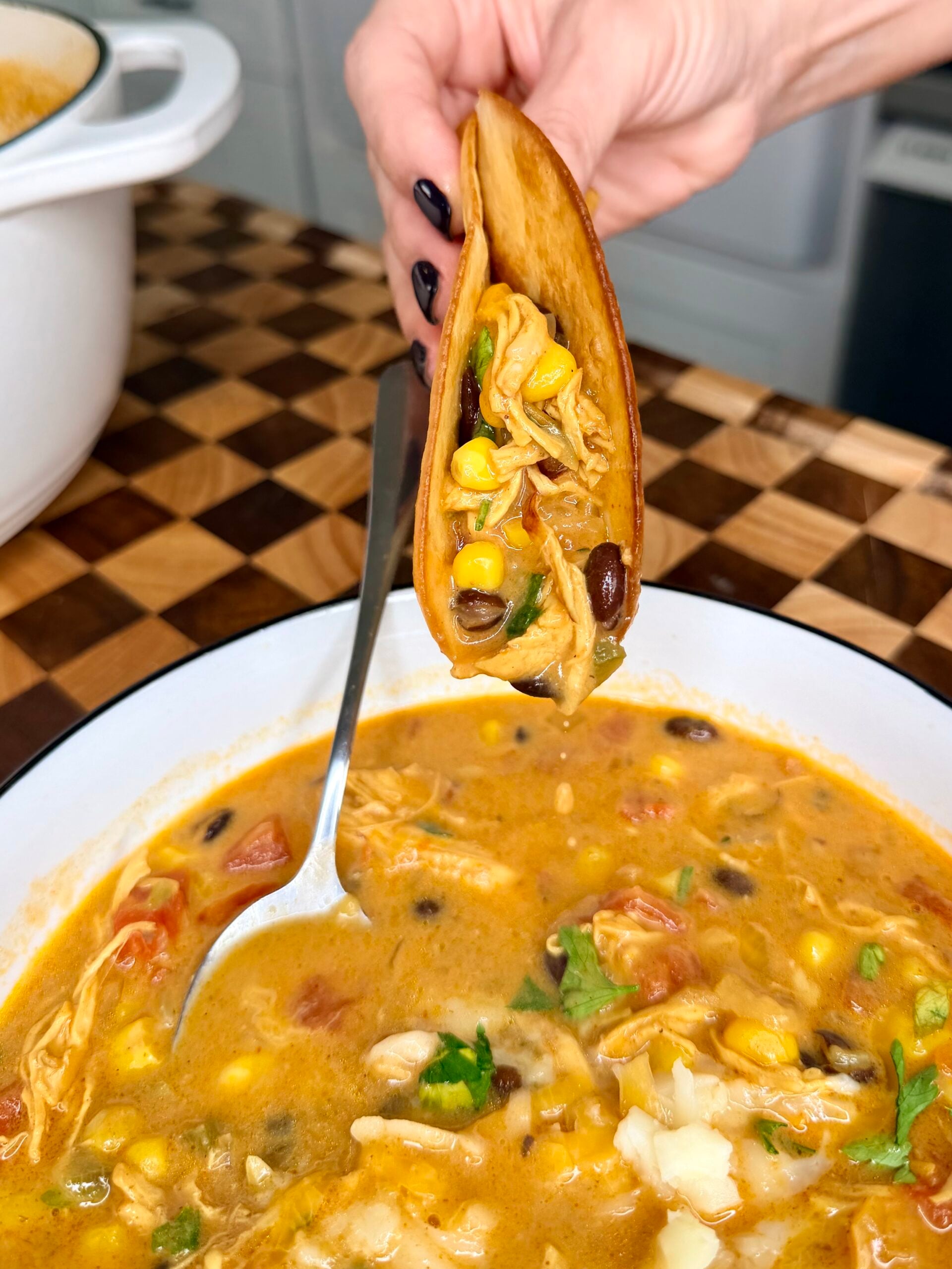 A hand holds a crispy tortilla shell filled with chicken, corn, and black beans above a bowl of chicken tortilla soup.