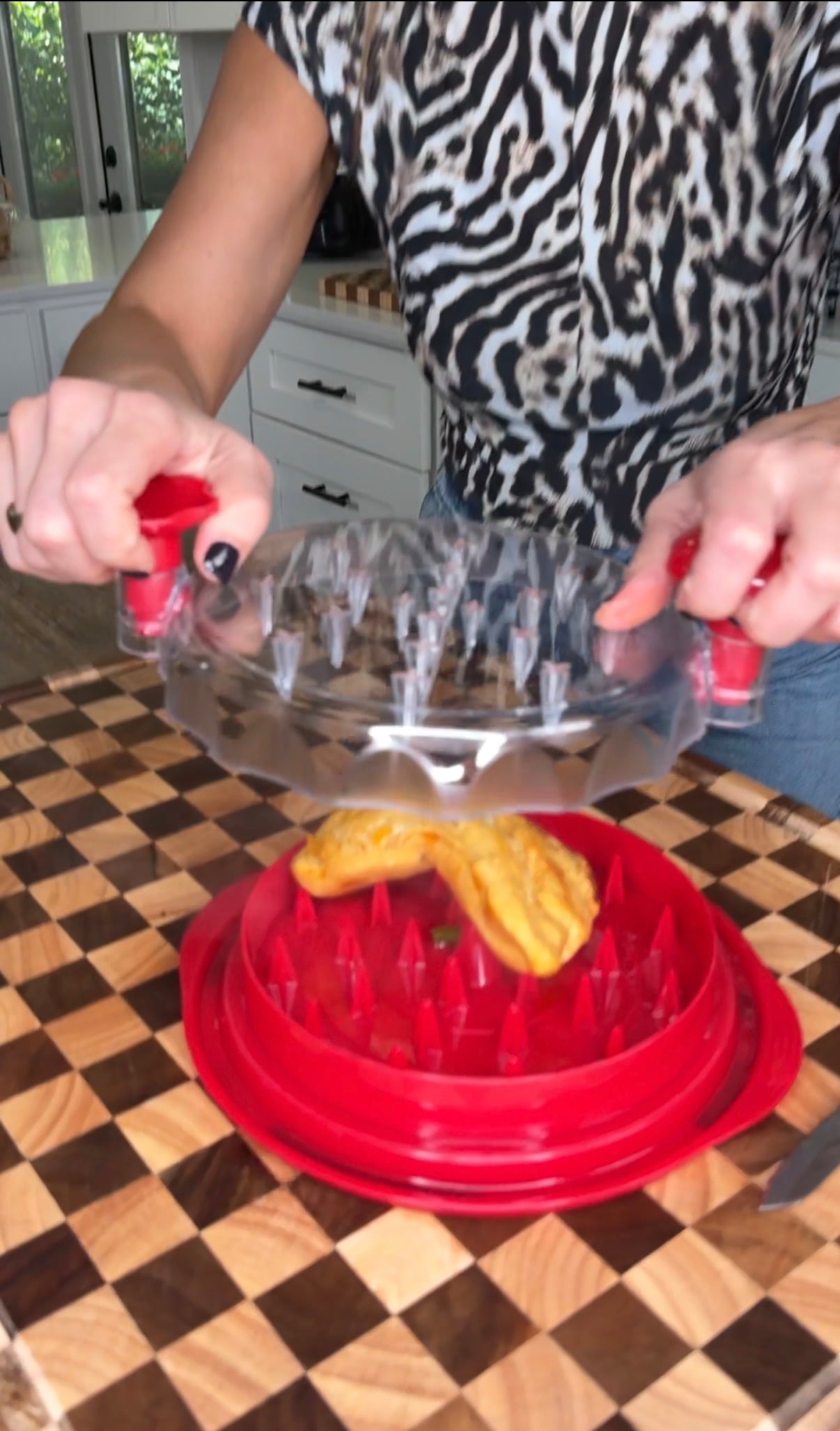 A person presses a clear plastic meat tenderizer tool down onto a piece of cooked chicken resting on a red base.