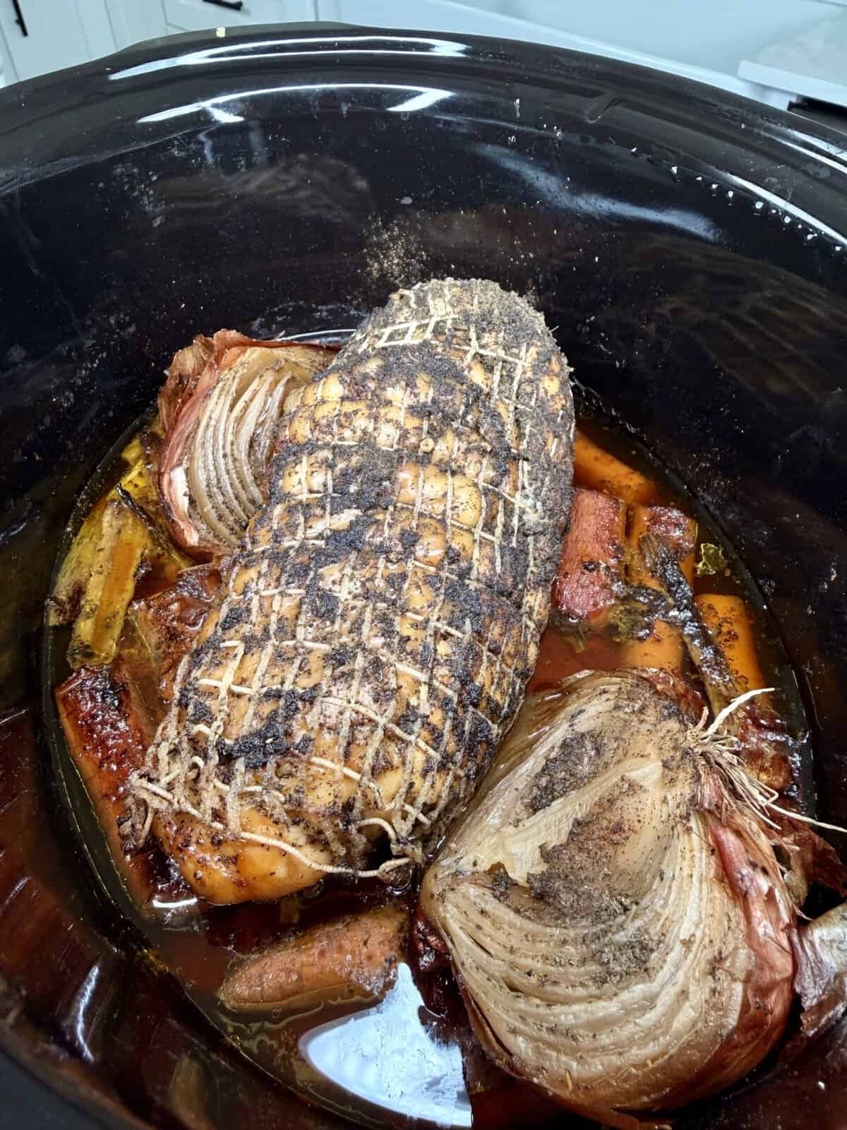 A seasoned roast in a netting wrap cooking in a slow cooker with halved onions and carrots.