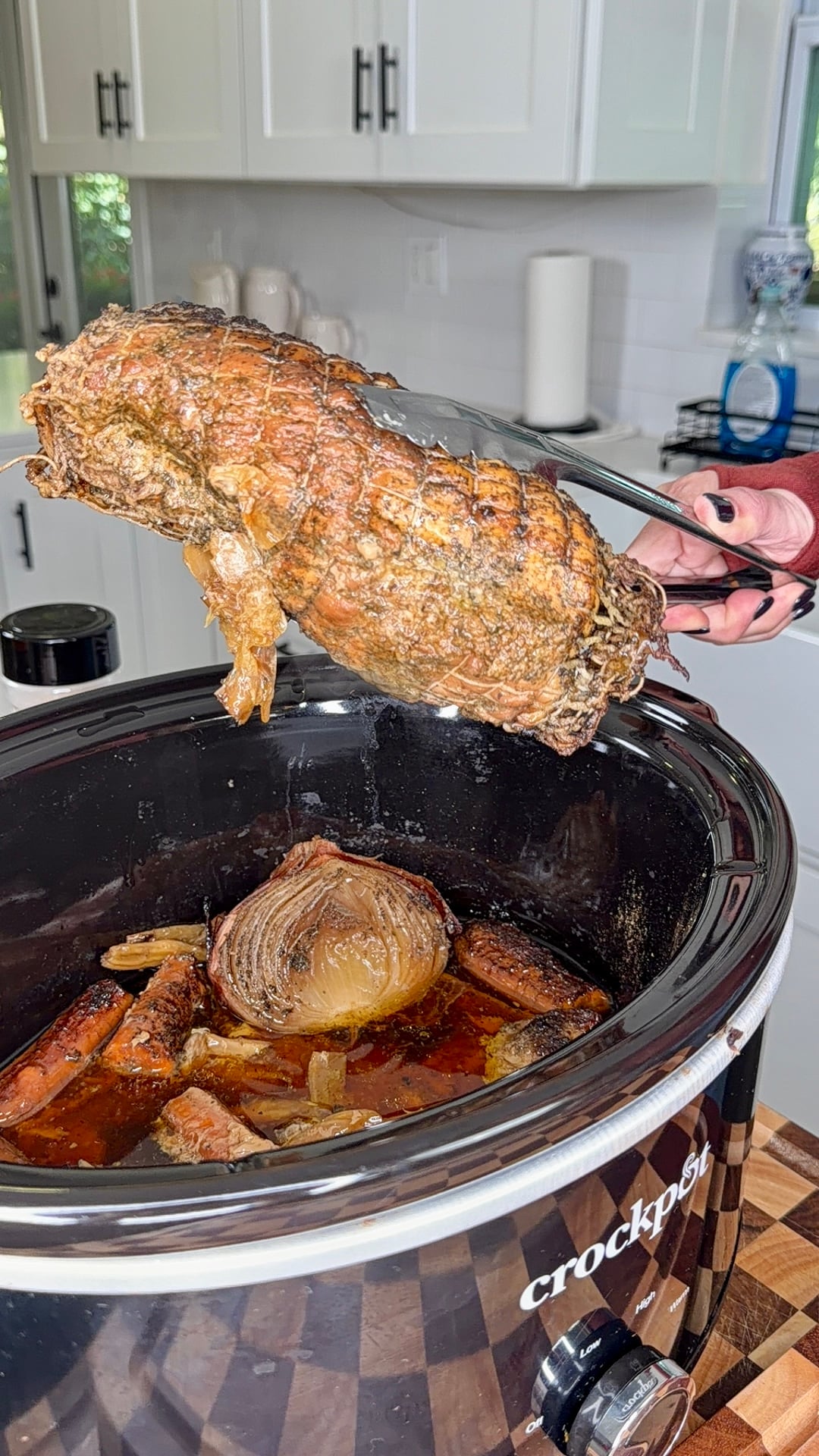 A hand uses tongs to lift a cooked, seasoned roast from a slow cooker containing onion and carrots in liquid.