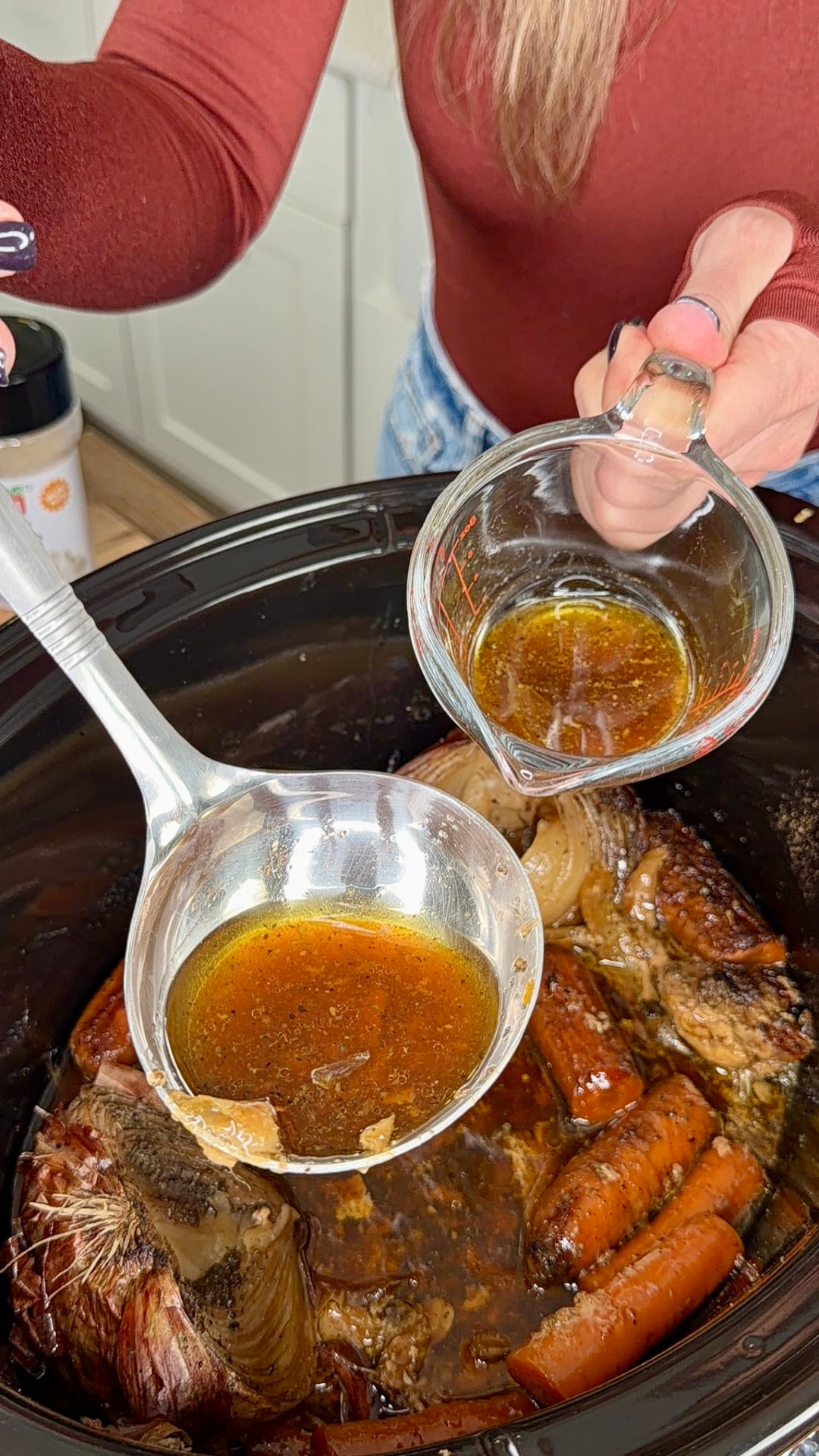 Hands pouring liquid from a glass measuring cup into a metal ladle held over a slow cooker containing a roast and carrots.