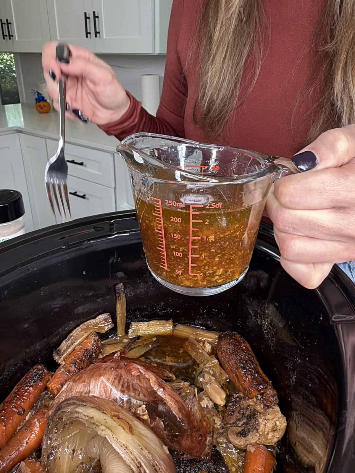 A person pours a brown liquid mixture from a measuring cup into a slow cooker containing roasted carrots and onions.