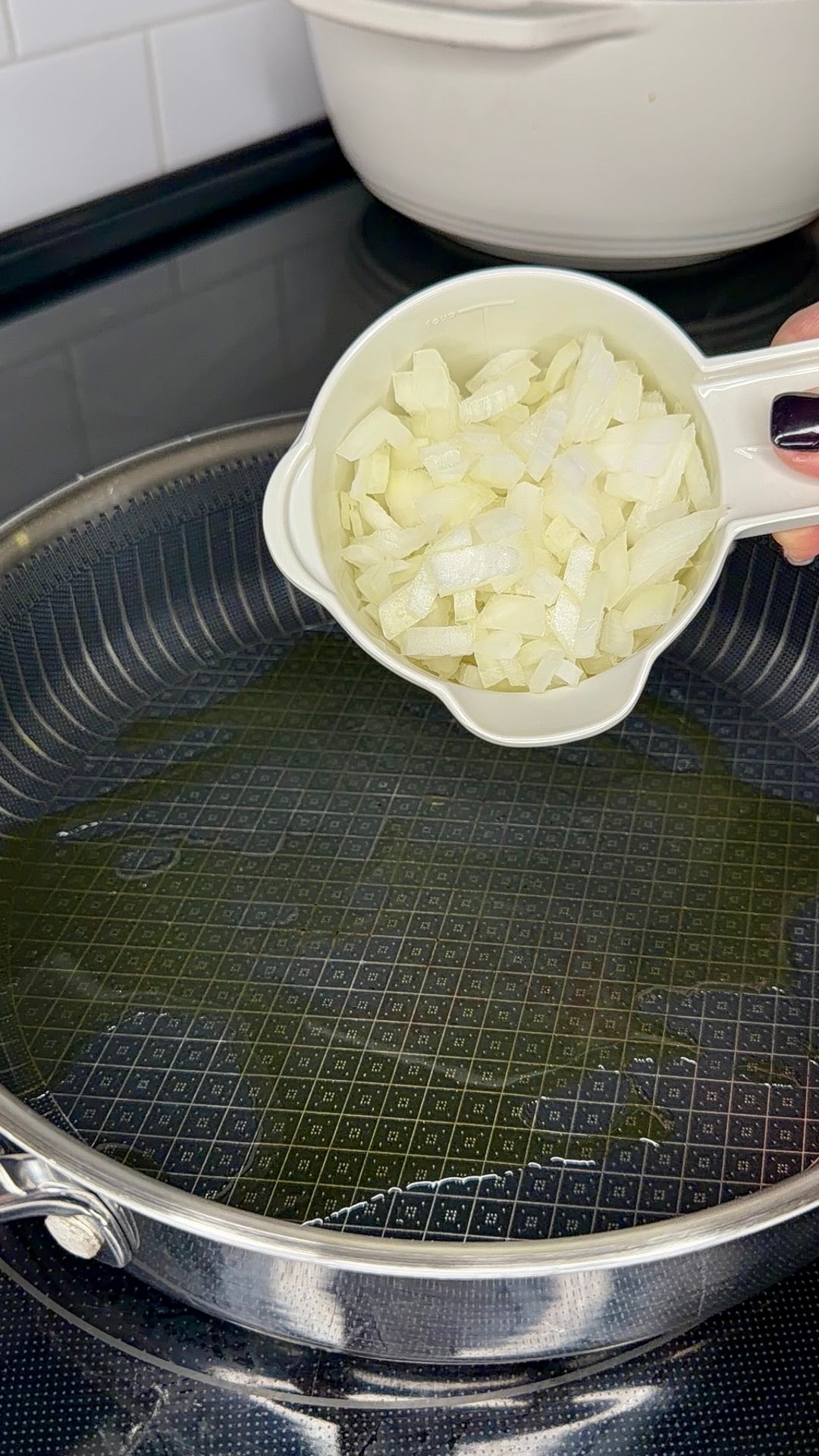 A hand pours a measuring cup filled with diced white onions into a frying pan with oil.