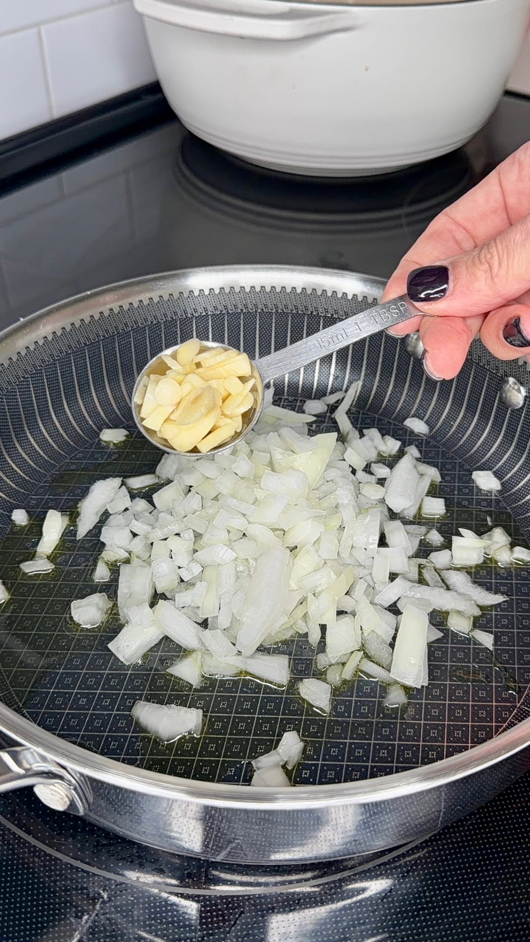 A hand holds a tablespoon filled with chopped garlic over a skillet containing diced onions sautéing in oil.