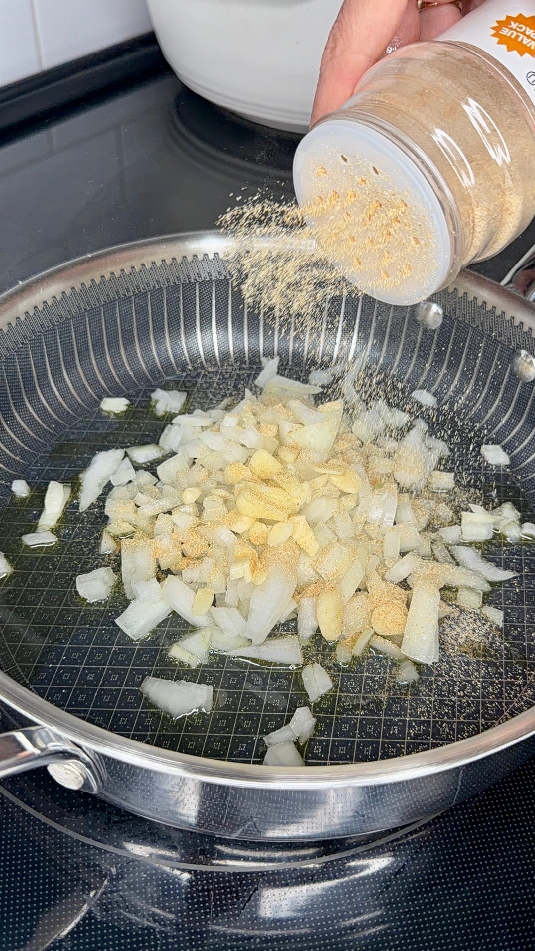 Hand holding a spice jar sprinkles powder over chopped onions and garlic in a metal frying pan.