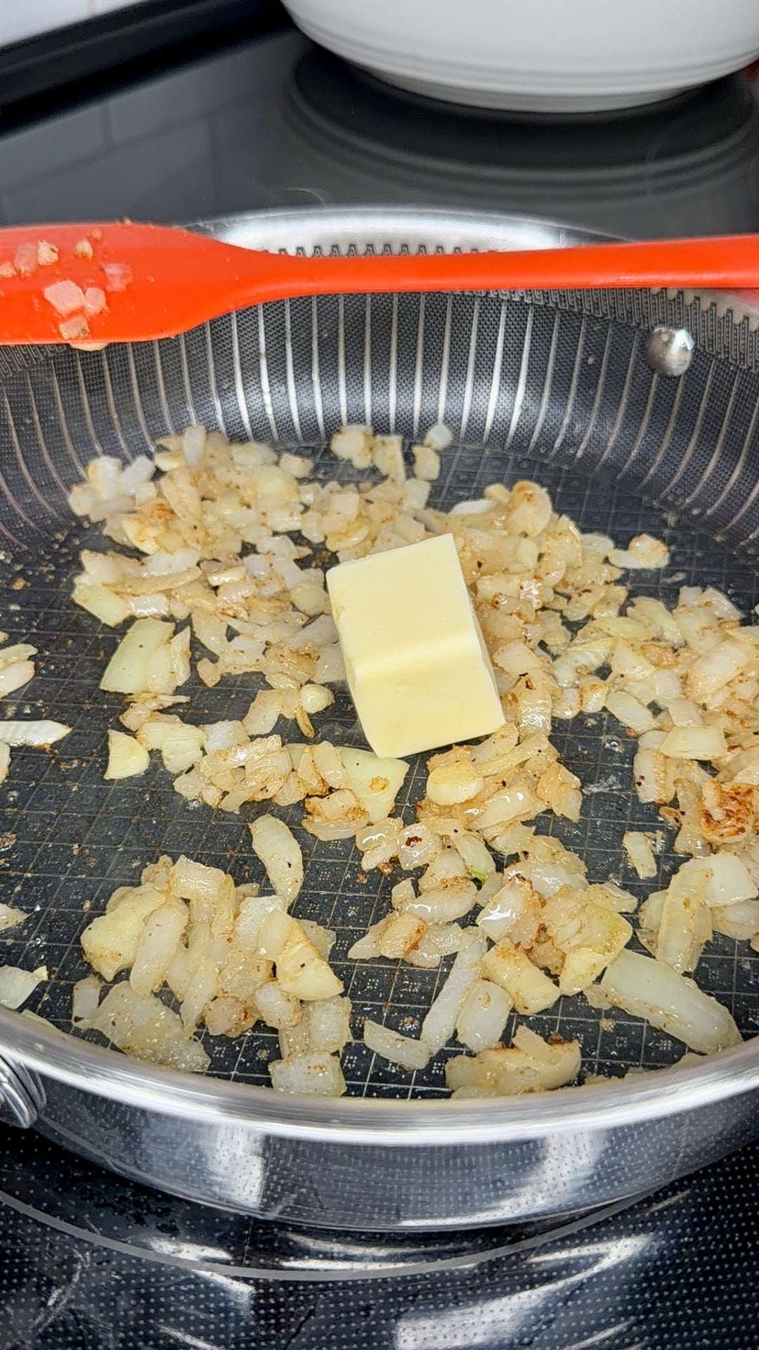 A pat of butter melting into sautéed onions in a frying pan with an orange spatula resting above.