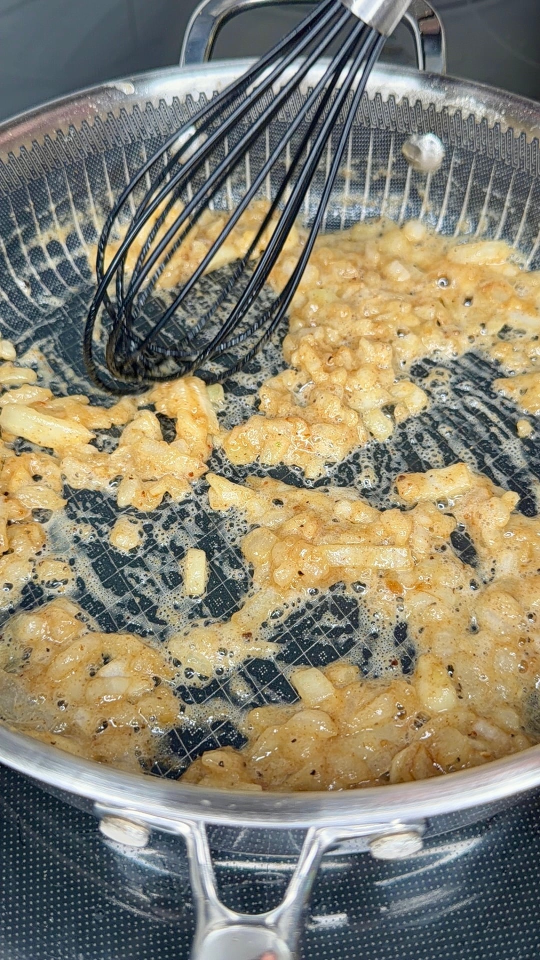 A wire whisk stirs diced onions sautéing in a stainless steel skillet.