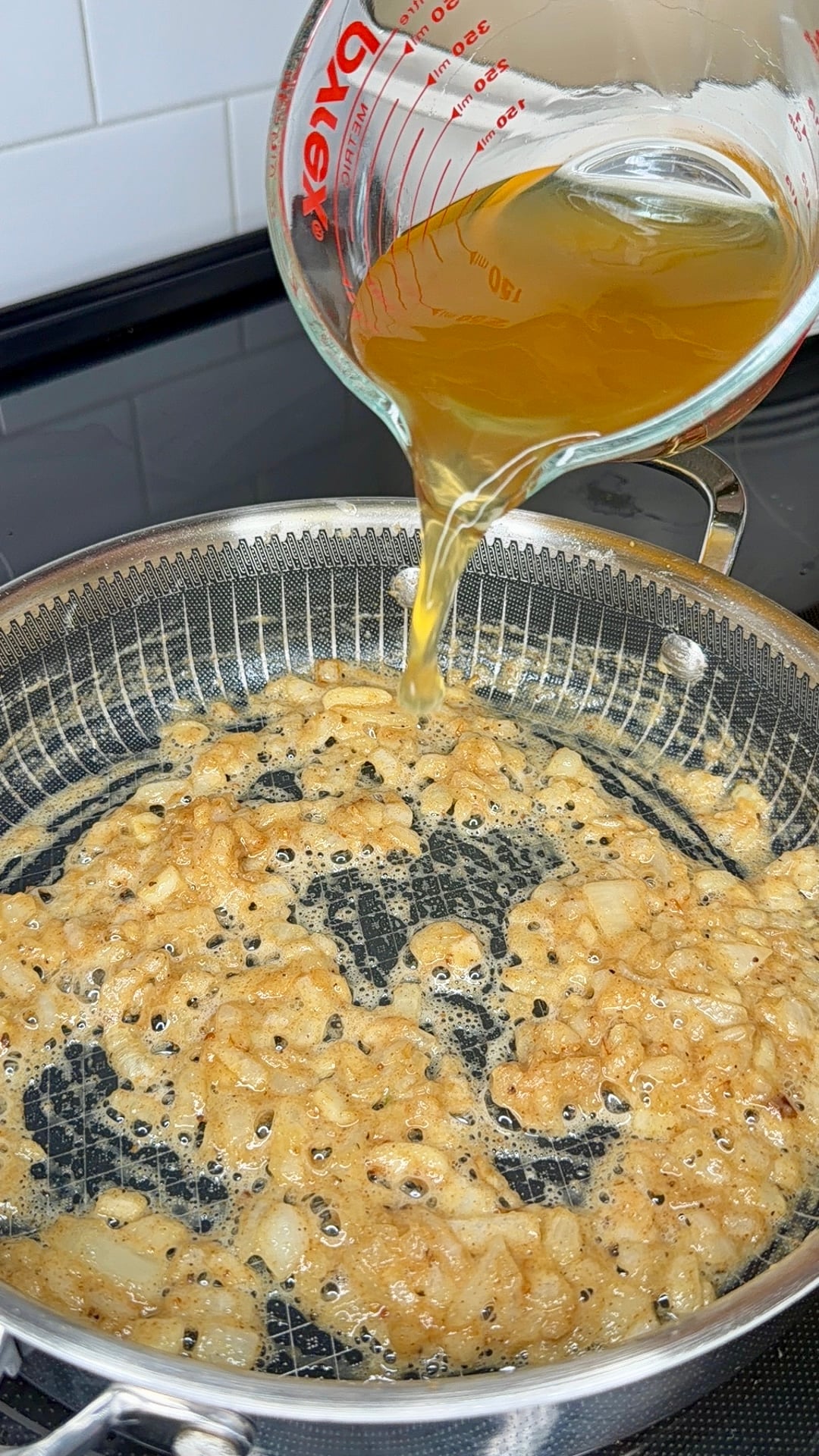 Broth being poured from a Pyrex measuring cup into a pan of sautéed onions.