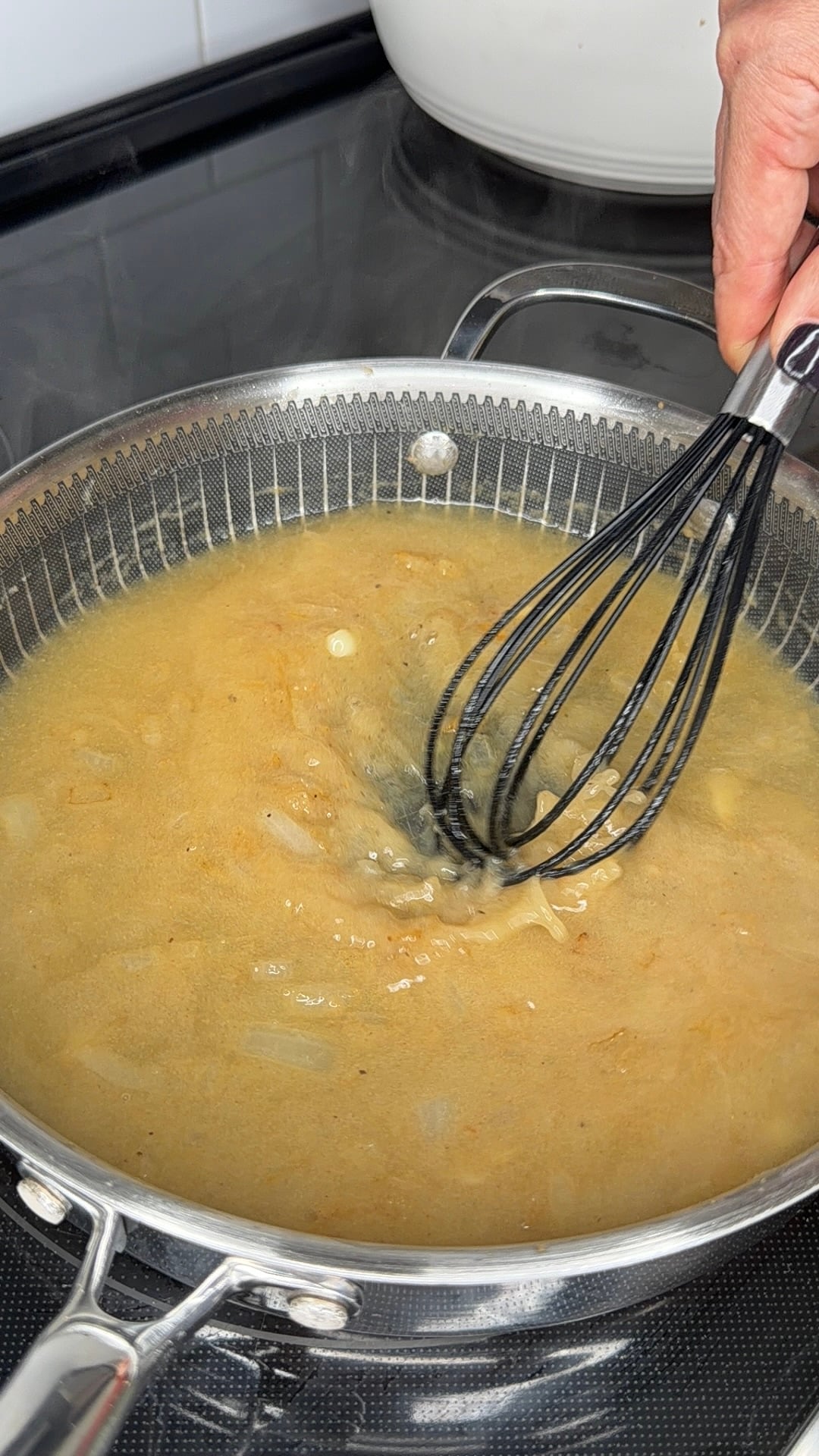 A hand uses a black whisk to stir a golden-brown liquid with visible pieces of onion in a stainless steel skillet on a stovetop.