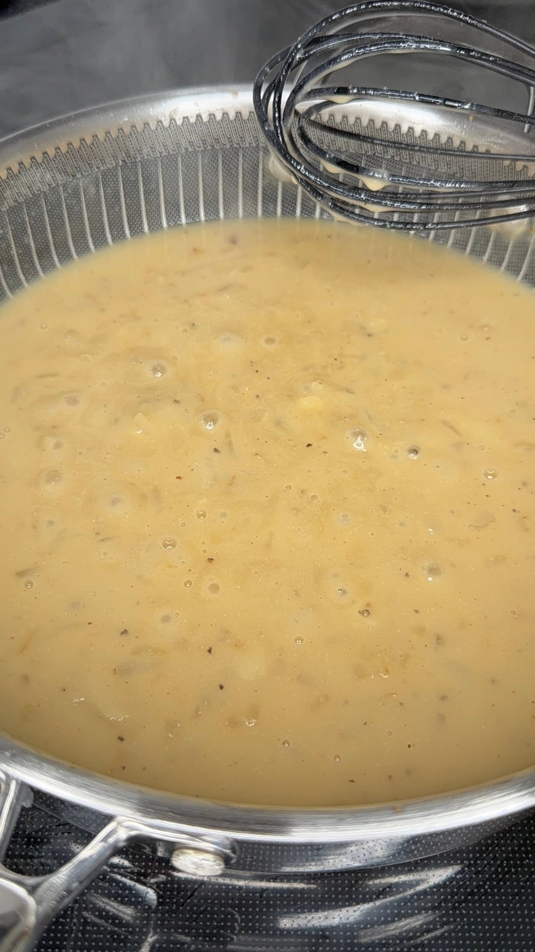 A wire whisk rests in a pot of simmering, creamy soup with visible herbs and spices.