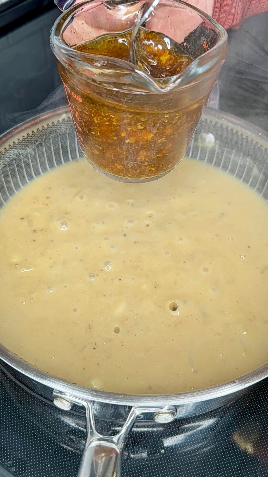 A hand pours a dark, textured sauce from a glass measuring cup into a pan of simmering cream-colored soup.