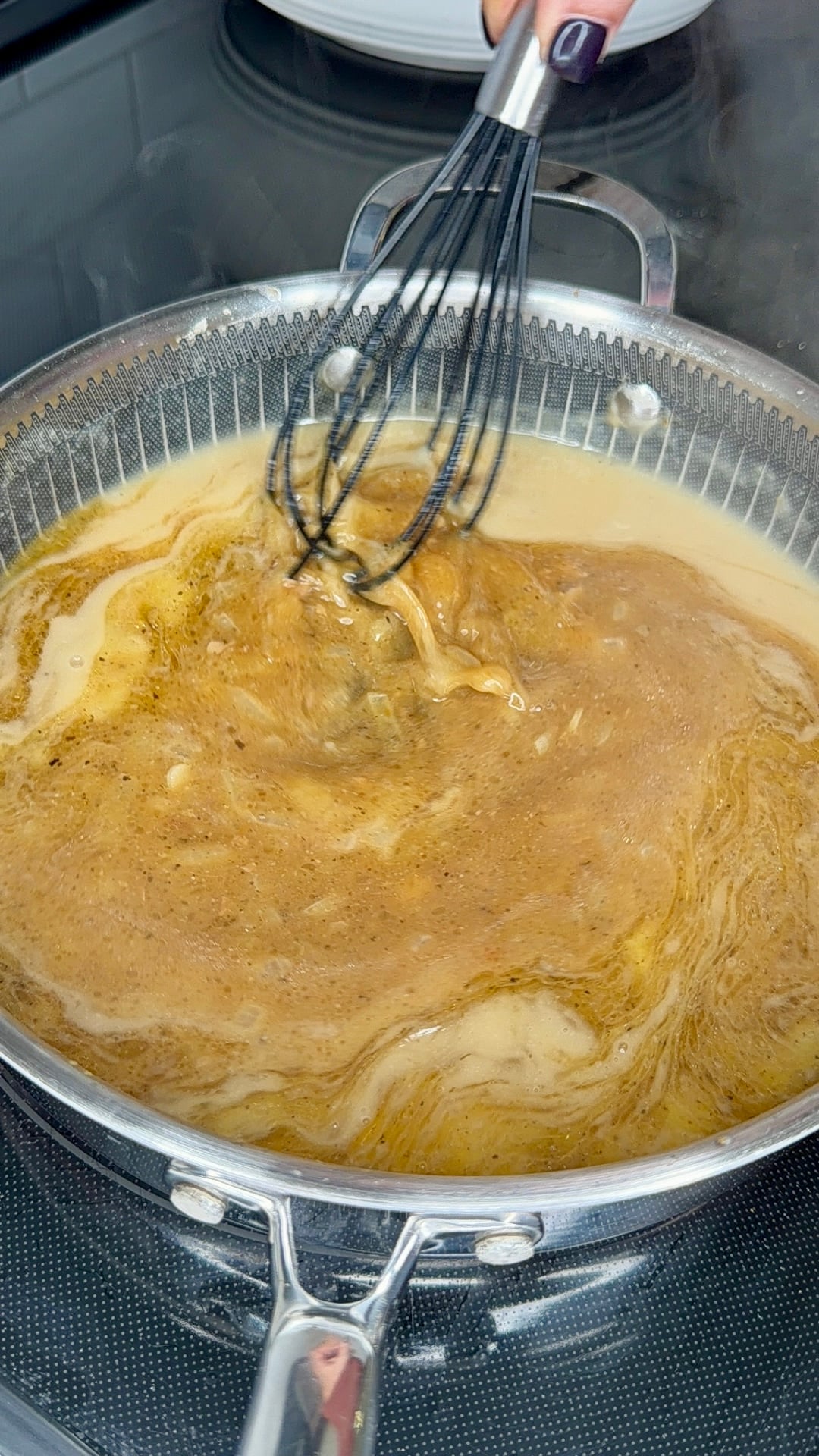 A hand uses a wire whisk to stir a golden-brown liquid in a stainless steel pan on a stovetop.