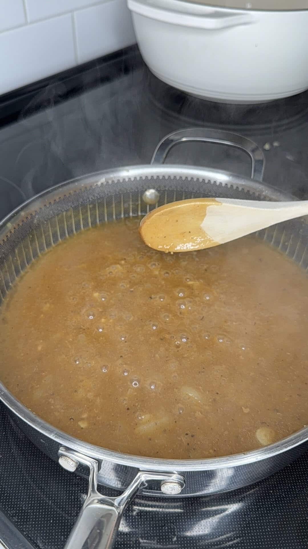 A wooden spoon lifts steaming brown sauce from a stainless steel pan on a stovetop.