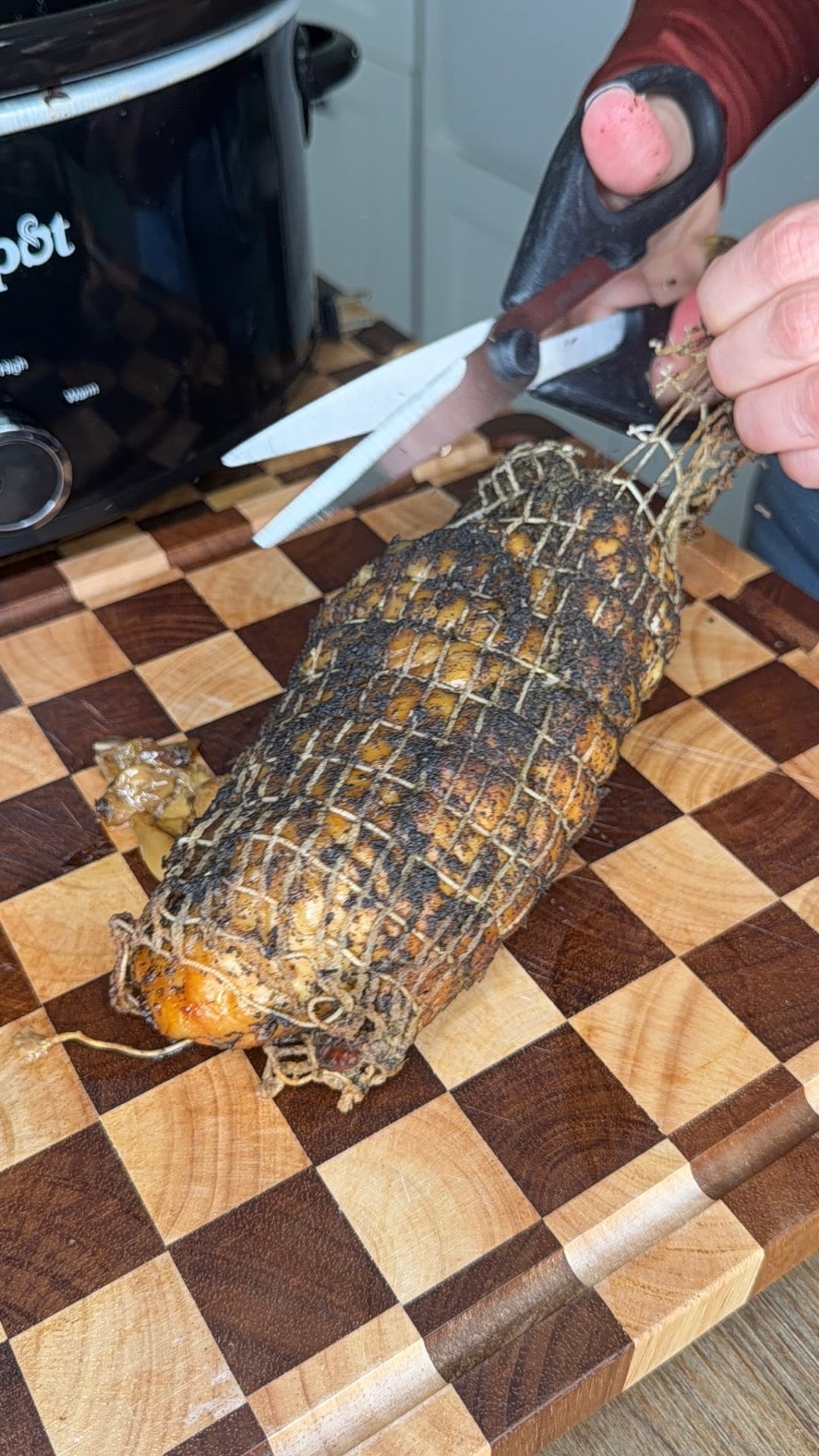 Hands use kitchen shears to cut the netting off a cooked, seasoned roast on a checkerboard cutting board.