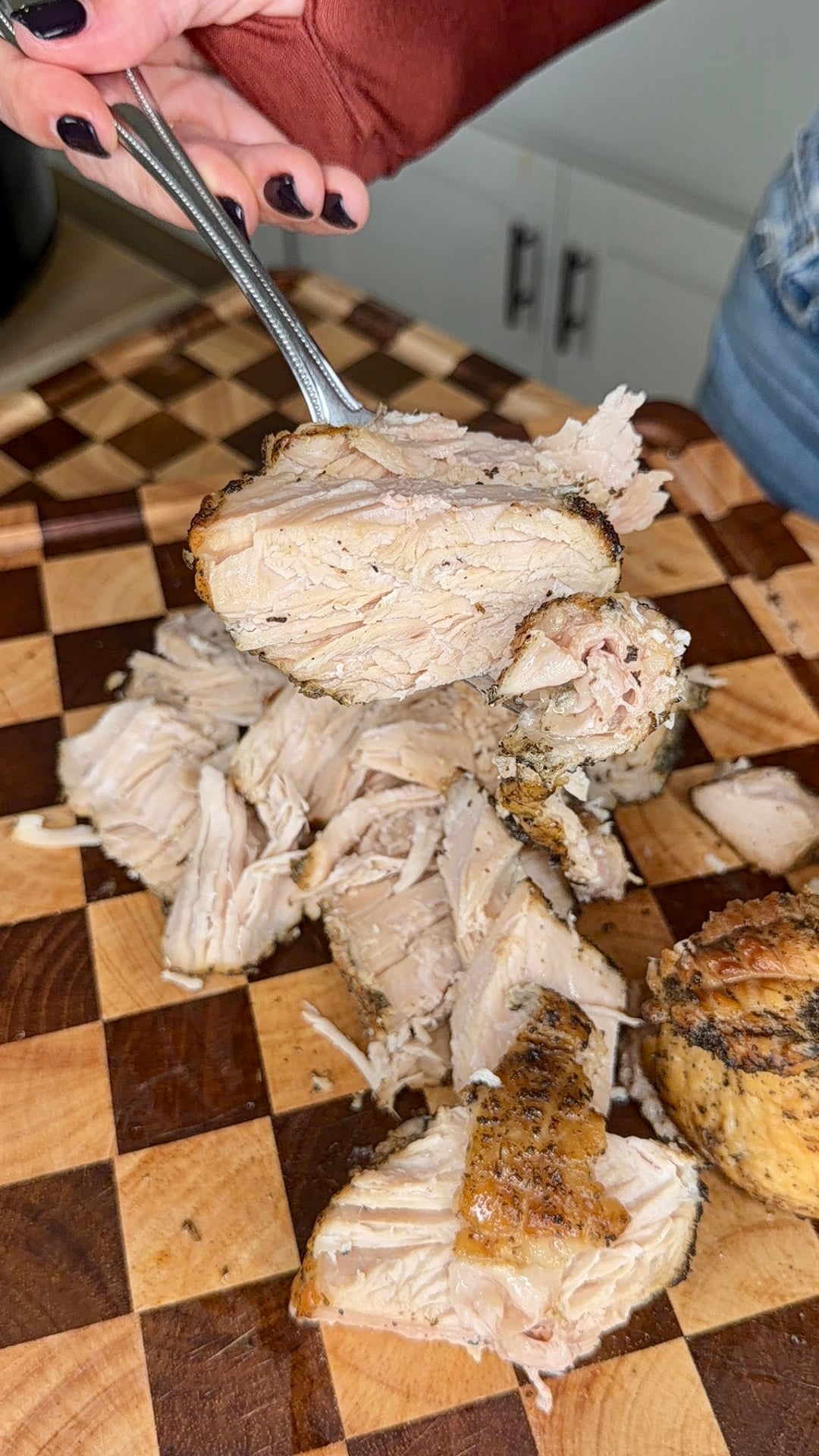 A fork lifts a slice of roasted chicken breast above a pile of shredded cooked chicken on a checkerboard cutting board.