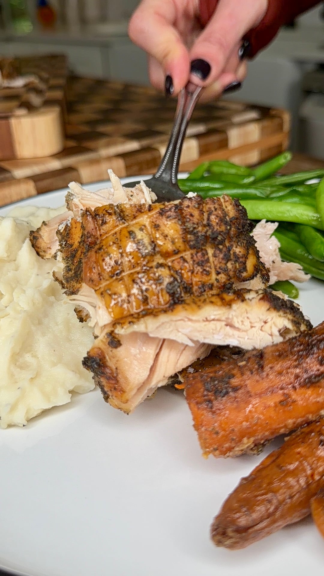A fork lifts a piece of seasoned, roasted turkey from a plate containing mashed potatoes, green beans, and roasted carrots.