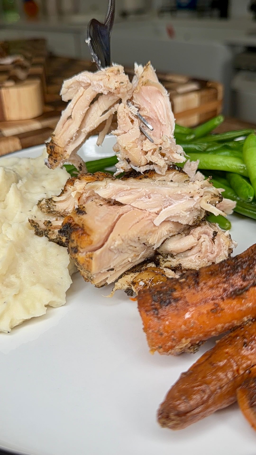 A fork holds a piece of shredded roasted chicken above a plate with mashed potatoes, green beans, and roasted carrots.