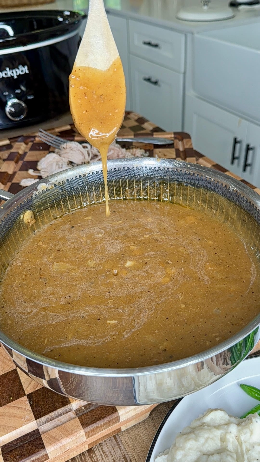 A wooden spoon dripping brown gravy over a large stainless steel pot filled with the same sauce.