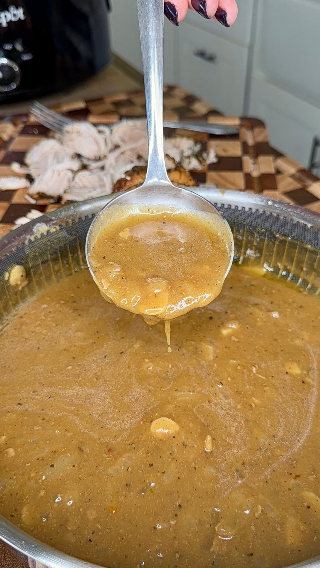 A metal ladle held over a pot of brown gravy, with shredded meat in the background.