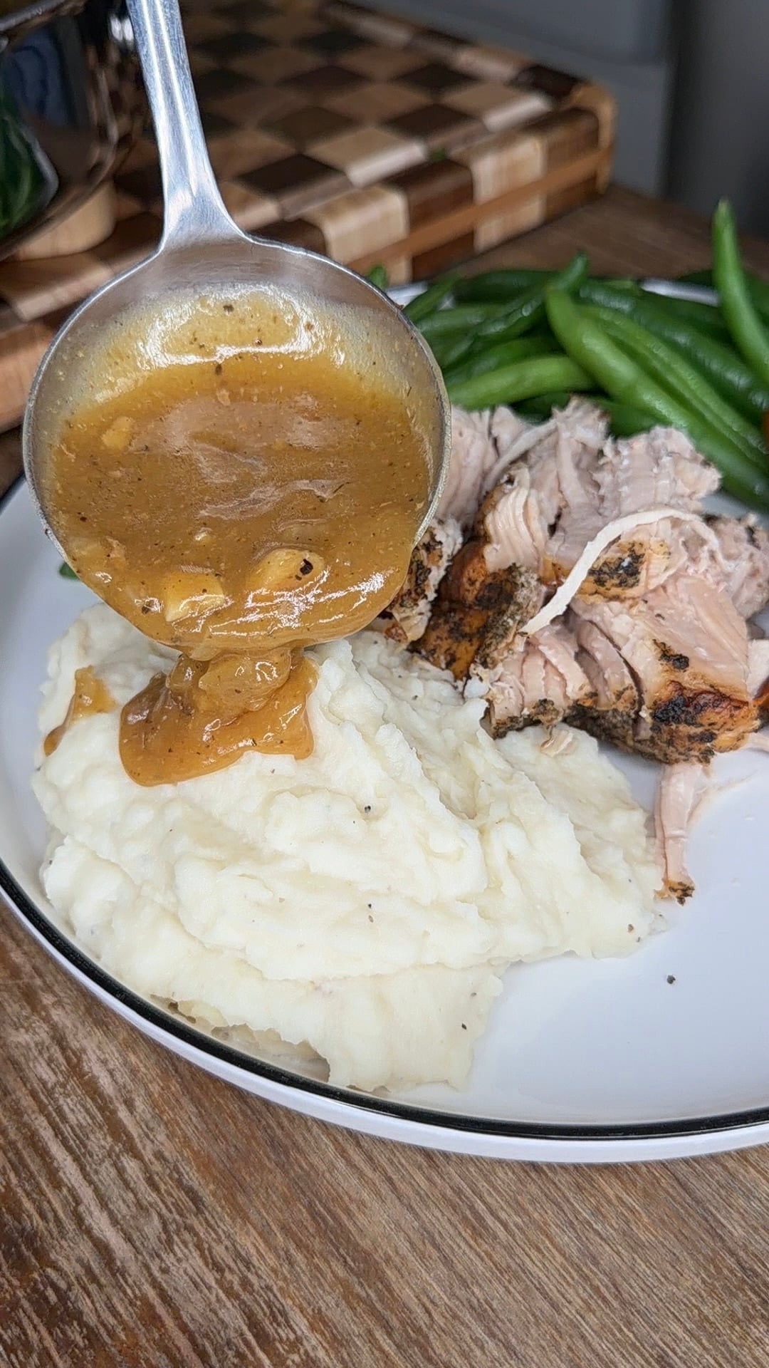 A ladle pours brown gravy over a mound of mashed potatoes next to shredded meat and green beans on a plate.