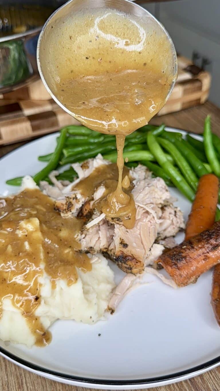 A ladle pours gravy over a plate of shredded chicken, mashed potatoes, green beans, and roasted carrots.