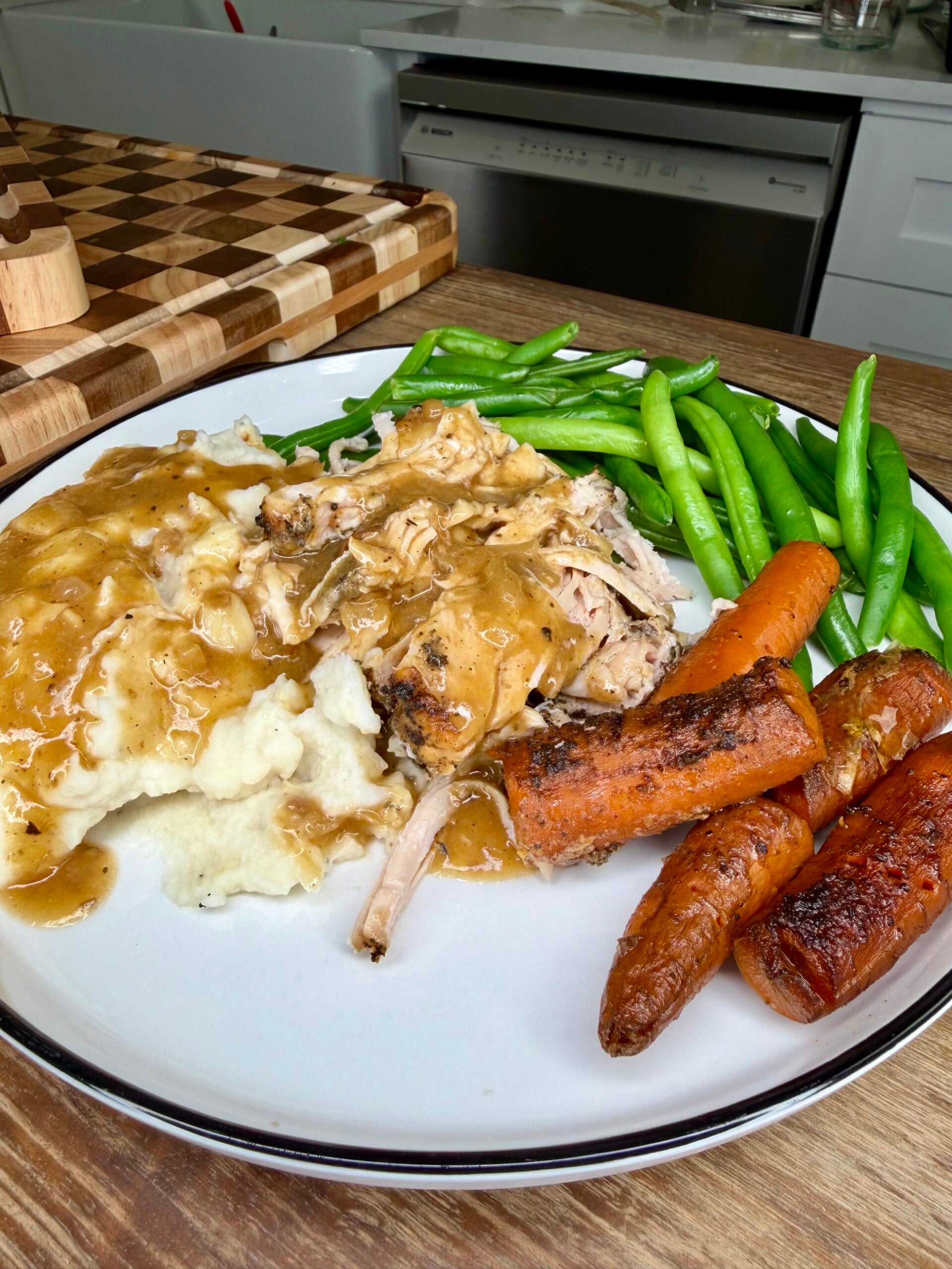 A plate of roasted chicken and mashed potatoes covered in gravy, served with green beans and roasted carrots.