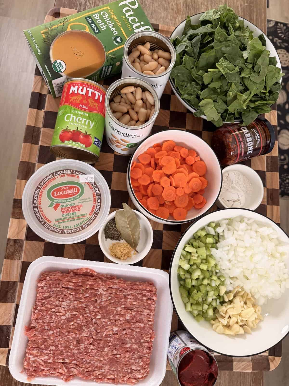 Ingredients for a recipe arranged on a cutting board, including ground meat, spinach, carrots, beans, onion, celery, garlic, chicken broth, canned tomatoes, tomato paste, cheese, and spices.