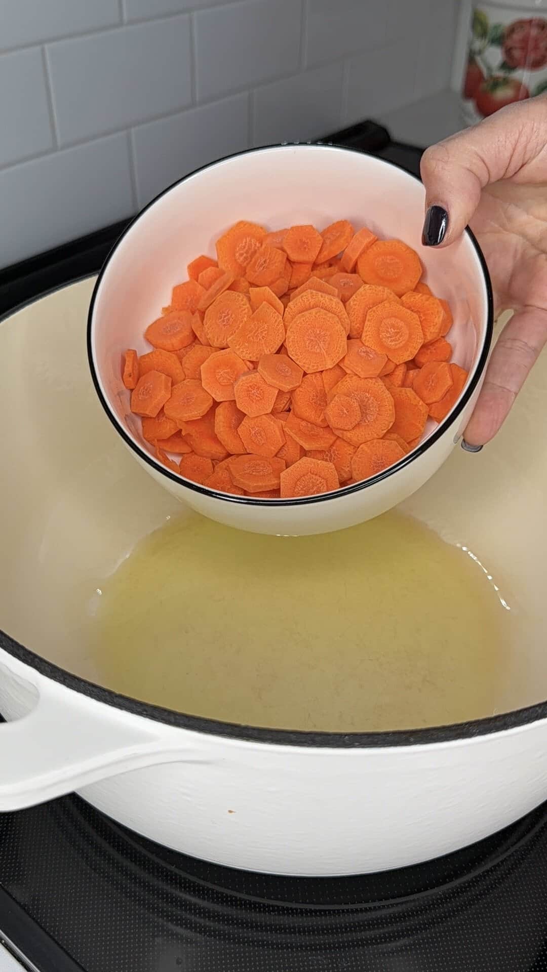 A hand holds a bowl of sliced carrots over a pot containing hot oil.