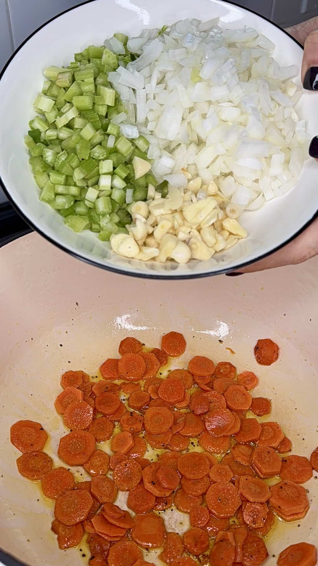 A person holds a bowl of diced celery, onions, and garlic over a pan containing sliced, sautéing carrots.