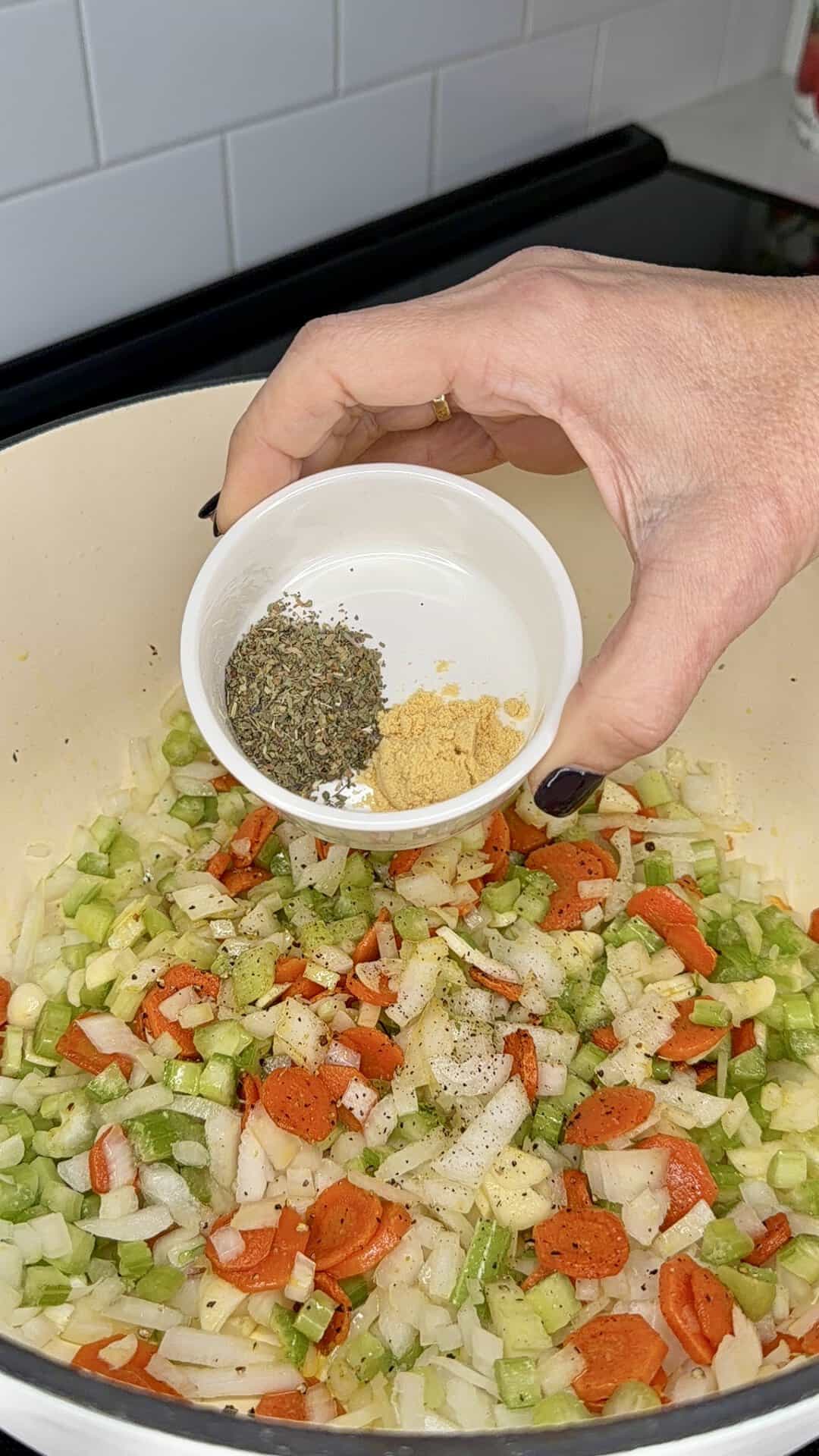 A hand holds a small white bowl over a pot of sautéing carrots, celery, and onions, pouring in dried herbs and ground spices.