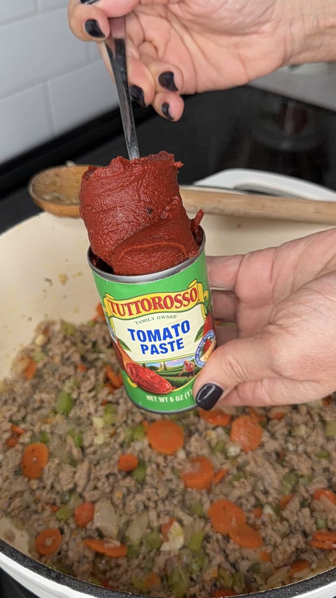 A hand holds a can of Tuttorosso tomato paste over a pot of cooked ground meat and vegetables.