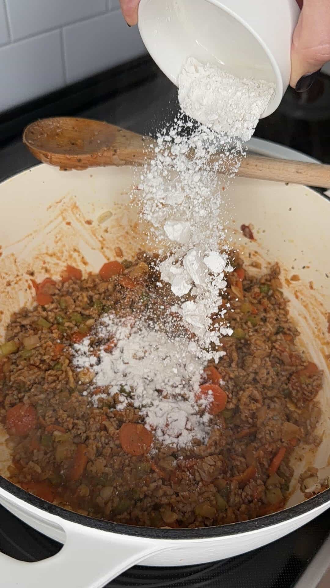 A hand pours flour from a white bowl into a pot containing a mixture of cooked ground meat and chopped vegetables.