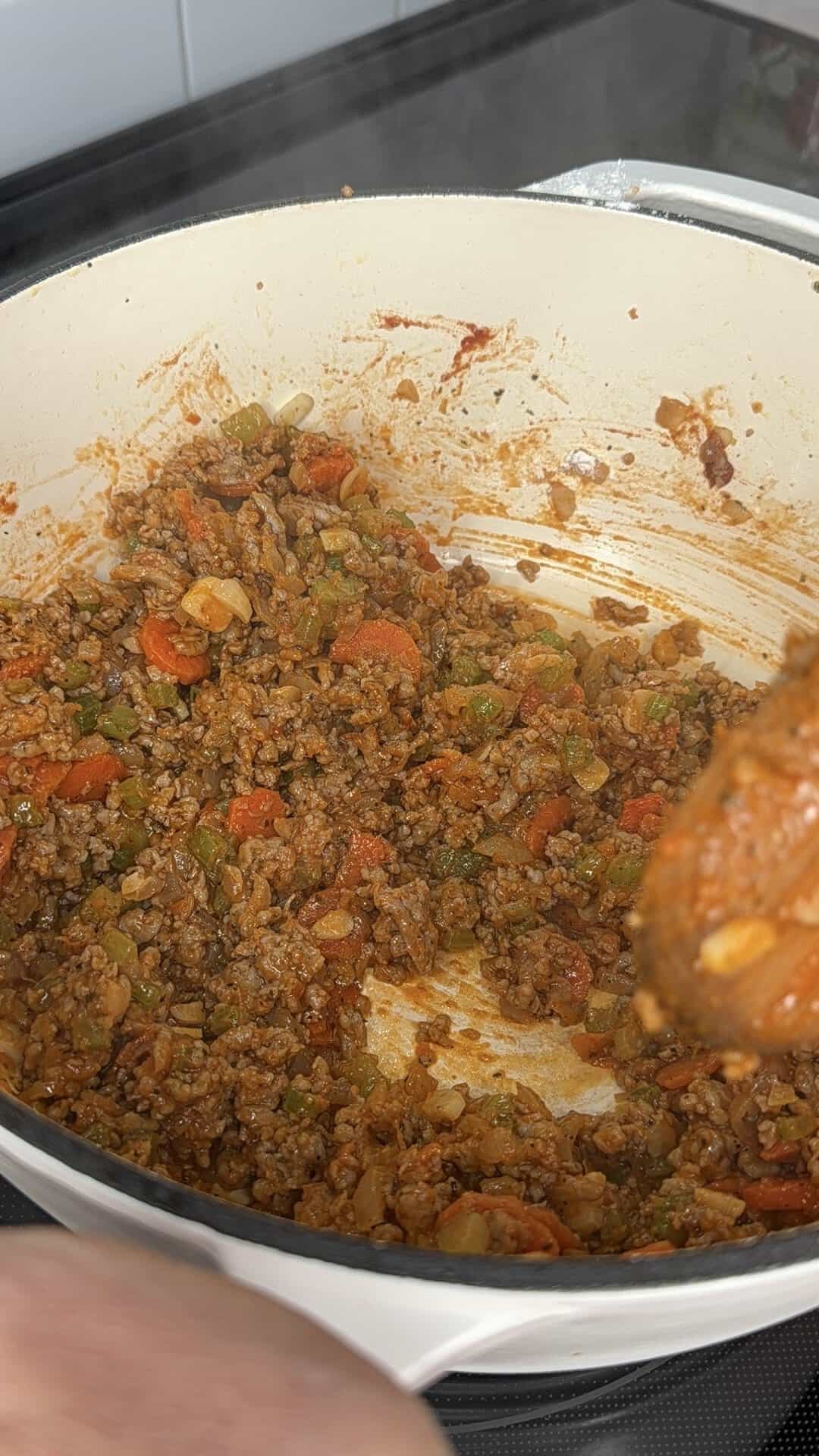Ground meat, diced carrots, and celery cooking in a white enameled pot.