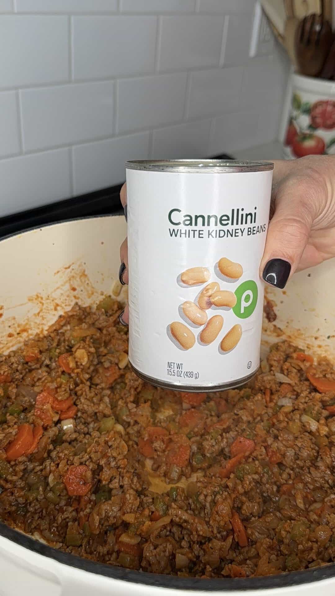 A hand holds a can of Cannellini white kidney beans over a pot of ground meat and vegetables cooking on a stovetop.