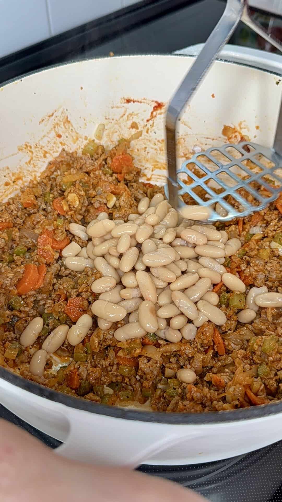 A hand-held masher presses white beans into a mixture of browned meat and vegetables cooking in a pot.