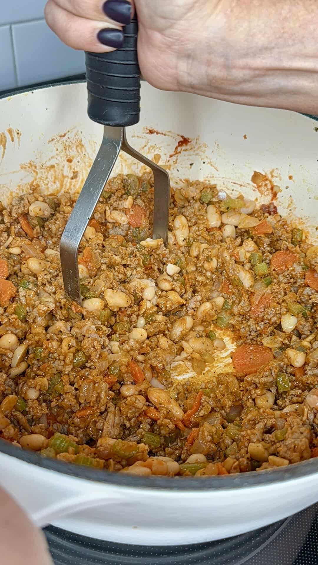 A hand uses a metal masher to combine ground meat, white beans, and chopped vegetables in a white pot.
